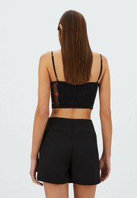 Vrouw met lang, steil bruin haar, gekleed in een zwart cropped top met bloemensluiting aan de zijkanten en hoge zwarte shorts, met haar rug naar ons gekeerd.