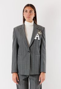 Blazer grigio a righe sottili su misura con due tasche, chiusura a un bottone e un patch ricamato con una "A" sul lato sinistro del petto.