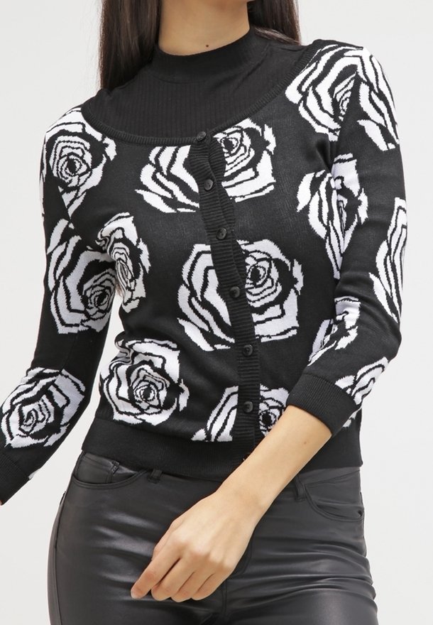 Femme portant un pull noir boutonné avec de grands motifs de roses blanches et un pantalon en cuir noir, main reposant près de la hanche.