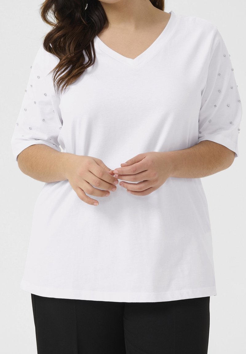 T-shirt en coton blanc avec un col en V et des manches courtes, orné de perles transparentes décoratives sur les épaules. Conception ample, texture lisse.