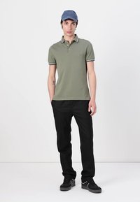 Ελιά πράσινο πόλο μπλουζάκι με λευκές λεπτομέρειες, μαύρο παντελόνι, μαύρα αθλητικά παπούτσια και μπλε καπέλο, σε φόντο λευκό.