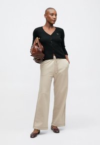 Sieviete ar noskūtu galvu, ģērbta melnā pogātā džemperī, bēšās platajās biksēs, brūnās kurpēs un turot rokās lielu brūnu ādas somu.