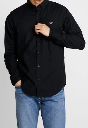 Hombre ajustándose una camisa negra de manga larga con botones y cuello, con un logotipo bordado en el pecho, y vistiendo jeans azul claro sobre un fondo liso.