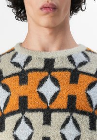 Strikkegenser med et geometrisk mønster i oransje, svart og lyseblått på en beige bakgrunn. Myk, lodden tekstur og rund hals.