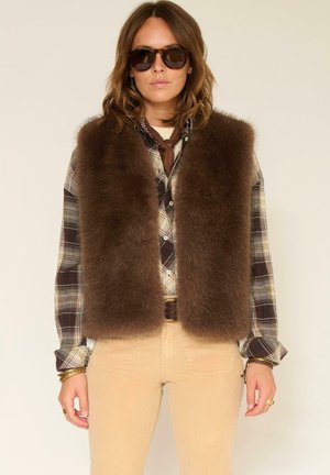 Gilet en fausse fourrure marron porté sur une chemise à carreaux, présentant un design court avec une texture douce. Les accessoires incluent des lunettes de soleil et un foulard marron autour du cou.