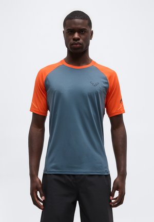 Jeune homme portant un maillot de sport à manches courtes avec des manches orange et un torse bleu, debout devant un fond blanc uni.