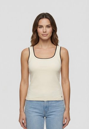 Donna con capelli castani di media lunghezza, indossa una canotta crema con bordi neri e jeans azzurri, di fronte a uno sfondo neutro.