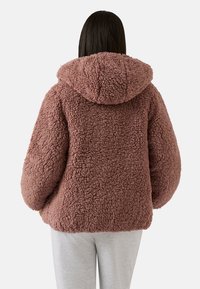 Brązowa, puszysta kurtka z kapturem wykonana z teksturowanej tkaniny, z dużymi rękawami i luźnym krojem. Widok z tyłu ukazuje zaokrąglony dół.