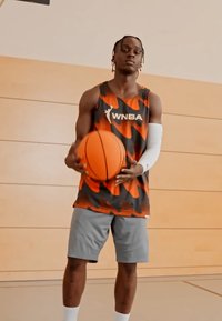 Jugor de baloncesto con una camiseta sin mangas de patrón camuflado en naranja y negro, pantalones cortos grises y una manga de brazo blanca, sosteniendo un balón de baloncesto naranja.
