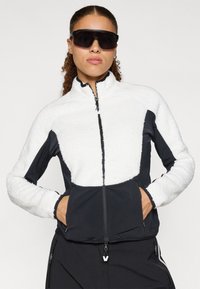 Fliisjakker, millel on valge tekstuuriga ülemine osa ja must sile alumine osa; omaduseks on lukuga esiosa, kõrge krae ja lukuga taskud.