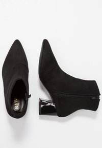 Botas de tobillo negras con puntera puntiaguda, hechas de ante, con cremallera lateral y tacones cuadrados brillantes. Textura suave con diseño minimalista.