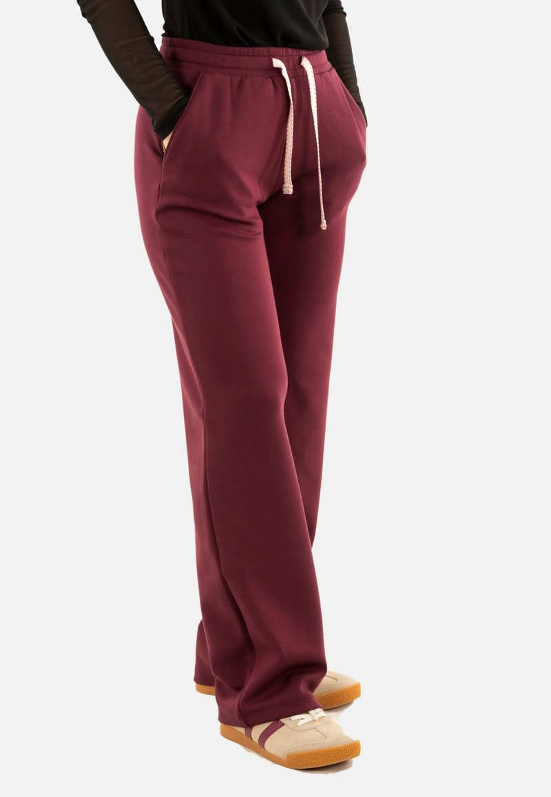 Pantalons de survêtement bordeaux avec un cordon de serrage, dotés de deux poches latérales et d'une coupe décontractée. Associés à des baskets beiges avec des semelles et des accents marron.