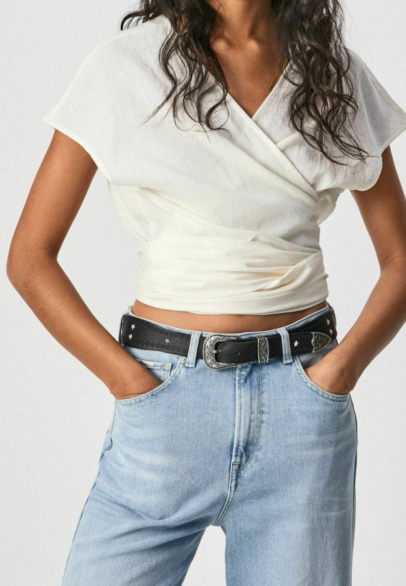 Koszulka w stylu wrap z kremowego materiału, z wysokim dekoltem i krótkimi rękawami. Nosić ją z jasnoniebieskimi spodniami w stylu high-waisted i czarnym dekoracyjnym paskiem.