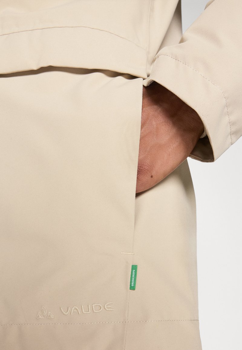 Lichtbeige Vaude-jack met een zak met een hand, textuurstof, geborduurd logo en een groene tag die eco-vriendelijkheid aangeeft.