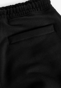 Pantalones de chándal negros con una cinturilla elástica y un bolsillo trasero con una solapa rectangular. La textura de la tela es suave y mate.