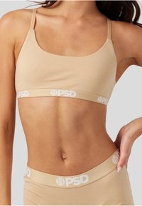 Beige bh med tynne stropper og bred merket bånd med "PSD" i hvit. Jevn tekstur og tettsittende design.