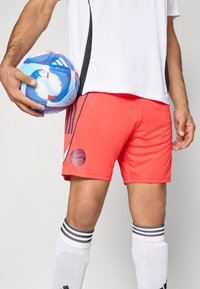 Červené športové šortky s logom FC Bayern Mníchov, v kombinácii s bielymi športovými ponožkami s čiernymi pruhmi a modrou futbalovou loptou s vzorom.