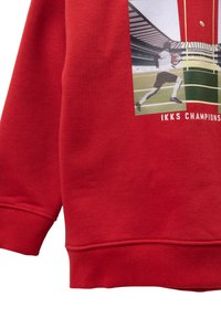Sudadera roja hecha de tejido suave que presenta una impresión gráfica de un jugador con el texto "IKKS CHAMPIONS" debajo. Puños y dobladillo de canalé.