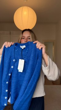 Μπλε ζακέτα με κουμπιά μπροστά, V-neck και μαλακή υφή. Ένδειξη τιμής συνημμένη, τοποθετημένη σε φόντο ανοιχτού χρώματος.