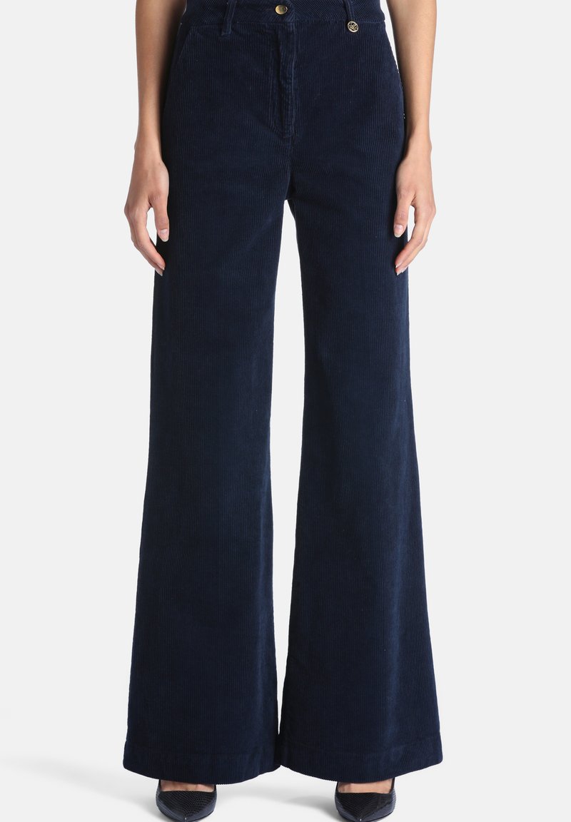 Pantalon large en velours côtelé bleu marine porté par une personne debout, les mains le long du corps, avec des chaussures noires à bout pointu visibles.