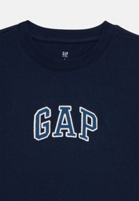 Tums zilgans kokvilnas krekls ar baltu un gaiši zilu izšūtu "GAP" logo priekšā, ar apaļu kakla izgriezumu un ribotu apkakli.