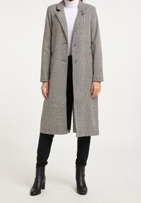 Cappotto lungo a scacchi grigio e bianco con vestibilità su misura, due bottoni e revers. Indossato sopra un dolcevita bianco e pantaloni neri.