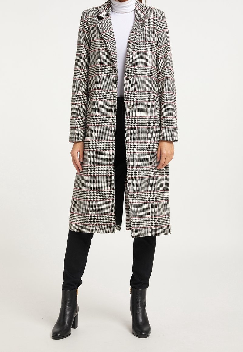 Cappotto lungo a scacchi grigio e bianco con vestibilità su misura, due bottoni e revers. Indossato sopra un dolcevita bianco e pantaloni neri.