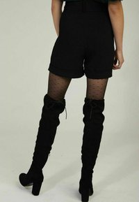 Shorts noirs sur mesure avec revers roulés, portés avec des collants noirs transparents à petits pois, et des bottes cuissardes en daim noir avec des accents de nœuds.