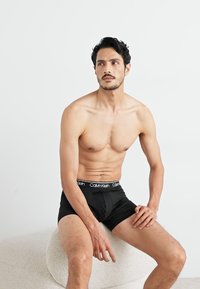 Crne Calvin Klein bokserice na muškom modelu; imaju glatku teksturu i elastičnu traku s brend logotipom. Model sjedi na svijetloj površini.