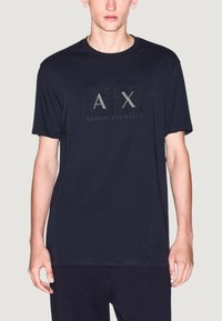 T-shirt en coton bleu marine à manches courtes, arborant un logo « AX » texturé et le texte « ARMANI EXCHANGE » dans un design rectangulaire.
