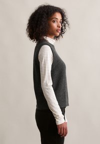 Grau ärmelloser Strickpullover, der über einem hellen karonierten Langarmshirt getragen wird, kombiniert mit dunklen Hosen. Die Textur wirkt weich und warm.