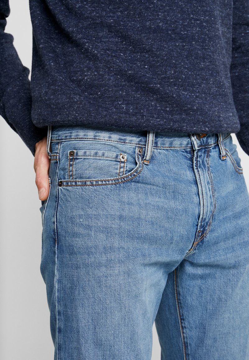 Verblasste blaue Jeans mit klassischem Fünf-Taschen-Design, Kupferbeschlägen und einem leichten Stretchanteil. Getragen mit einem dunklen, strukturierten Pullover.