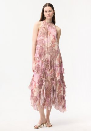 Giovane donna in piedi che indossa un vestito a balze senza maniche, rosa e beige con motivo paisley, e tacchi dorati aperti sulle dita dei piedi, su sfondo bianco.