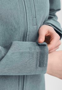 Pilkas užtrauktukas švarkas, pasižymintis tekstūruota medžiaga ir reguliuojamais rankogaliais su velcro užsegimu. Prekės ženklas "W PRO 800" matomas ant rankovės.