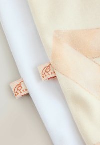 Divas audumi: viens balts un viens krēmkrāsas ar pasteļtoņu rakstu. Abiem ir mazas, iestrādātas etiķetes ar zīmola nosaukumu "Baker" rozā toņos.