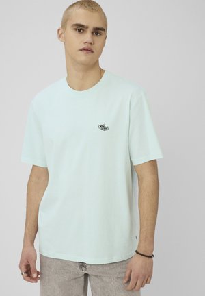 Jeune homme aux cheveux blonds courts, portant un t-shirt bleu clair et un jean gris clair, debout devant un fond gris uni.