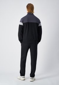 Completo sportivo nero e grigio con colletto a zip, caratterizzato da un design a blocchi di colore e polsini elasticizzati, abbinato a pantaloni coordinati e sneakers bianche.