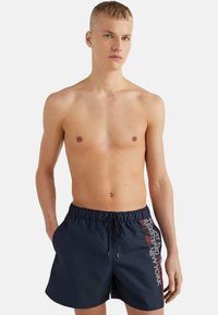 Bañador azul marino con cintura elástica y cordón. Presenta texto impreso en rojo a lo largo del costado. Fabricado con material ligero.