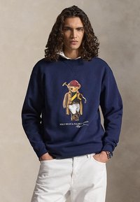 Moški s kodrastimi temnimi lasmi nosi temnomodro majico z medvedom v polo oblačilih, združeno z belimi hlačami, pozira na nevtralnem ozadju.