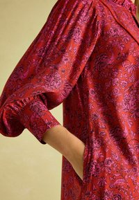 Red floral patterned dress with puff sleeves and gathered cuffs; pink and dark floral motifs; features side pockets.