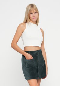 Młoda kobieta ubrana w białą bezrękawnikową bluzkę crop top i ciemnozieloną sztruksową spódnicę zapinaną na guziki z kieszeniami z przodu.
