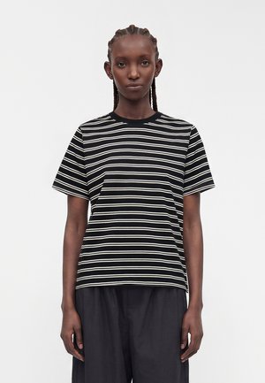 Personne aux cheveux tressés portant un t-shirt à manches courtes rayé horizontalement noir et blanc et un pantalon noir ample, se tenant devant un fond uni.