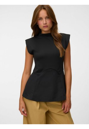 Femme aux longs cheveux bruns portant un haut noir sans manches avec épaules rembourrées et un pantalon beige sur un fond clair uni.