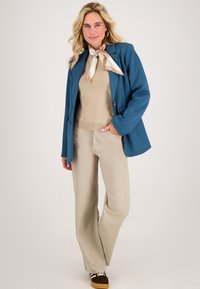 Teal blazer over beige top, gecombineerd met lichtbeige wijde broek. Accessoires zijn een crèmekleurige sjaal en bruine sneakers.