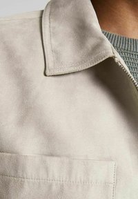 Ljusbeige mocka jacka med slagkrage, som har en framficka och en dragkedja. Texturen är mjuk med en slät finish.