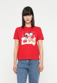 Woman wearing a red t-shirt with a holiday-themed print of Mickey and Minnie Mouse dressed as Santa Claus, paired with blue jeans.