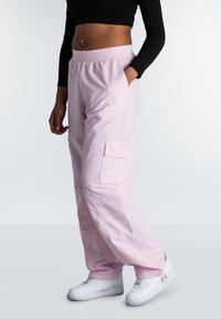 Roze cargobroek met zijzakken en een ontspannen pasvorm, voorzien van een elastische tailleband. Gepaard met witte sneakers op een neutrale achtergrond.