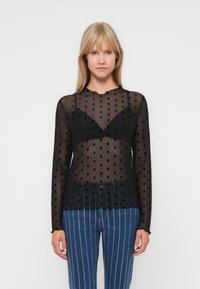 Transparante zwarte top met lange mouwen en gestructureerde stippen, met een scalloped halslijn, en een aansluitende zwarte bralette eronder, gecombineerd met gestreepte blauwe jeans.