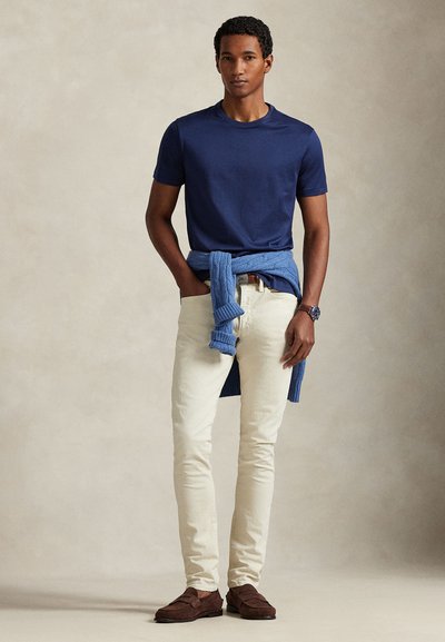 Άνδρας με μπλε ναυτικό μπλουζάκι, εκρού παντελόνι, καφέ μοκασίνια, μπλε πουλόβερ δεμένο γύρω από τη μέση και ρολόι χειρός, στέκεται μπροστά σε μπεζ φόντο.