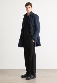 Cappotto trench navy sopra un maglione nero, abbinato a pantaloni neri e scarpe lucide, caratterizzato da un design strutturato e linee eleganti.